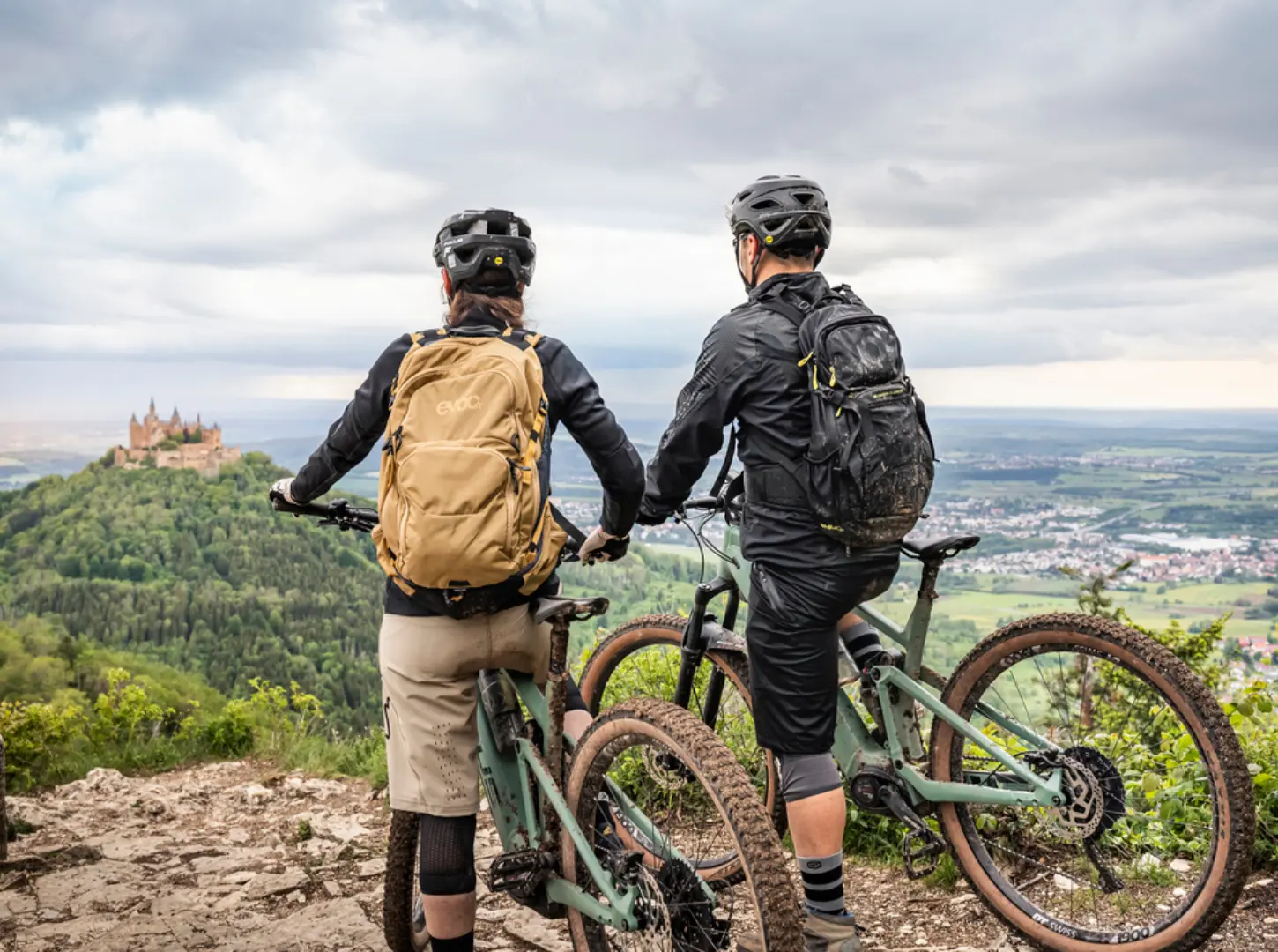 Eine Frau und ein Mann stehen mit ihren Focus Thron EBikes auf einem Berg mit Ausblick auf eine Burg